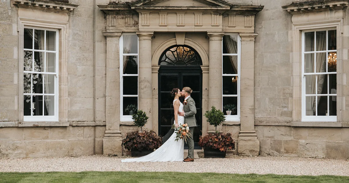 A newlywed couple kiss outside Bourton Hall, a grand stone building with tall windows and an arched doorway. The bride wears a white dress and holds a bouquet, while the groom dons a light suit. Two potted plants flank the entrance. © Aimee Lince Photography, Lincolnshire photographer