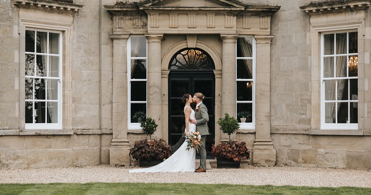 A newlywed couple kiss outside Bourton Hall, a grand stone building with tall windows and an arched doorway. The bride wears a white dress and holds a bouquet, while the groom dons a light suit. Two potted plants flank the entrance. © Aimee Lince Photography, Lincolnshire photographer