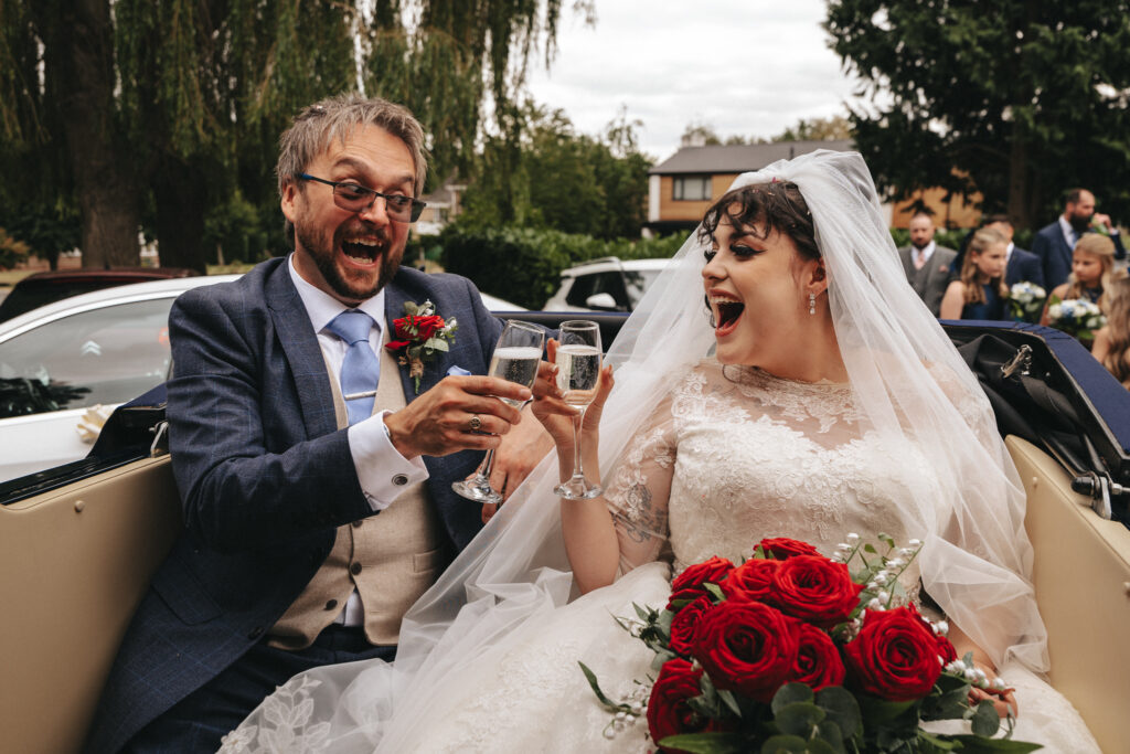 A joyful bride and groom sit in a convertible car, toasting with champagne glasses. The bride wears a white lace dress and veil, holding a bouquet of red roses, while the groom is in a blue suit and beige vest with a red boutonnière. Both appear to laugh heartily, surrounded by trees and guests. © Aimee Lince Photography