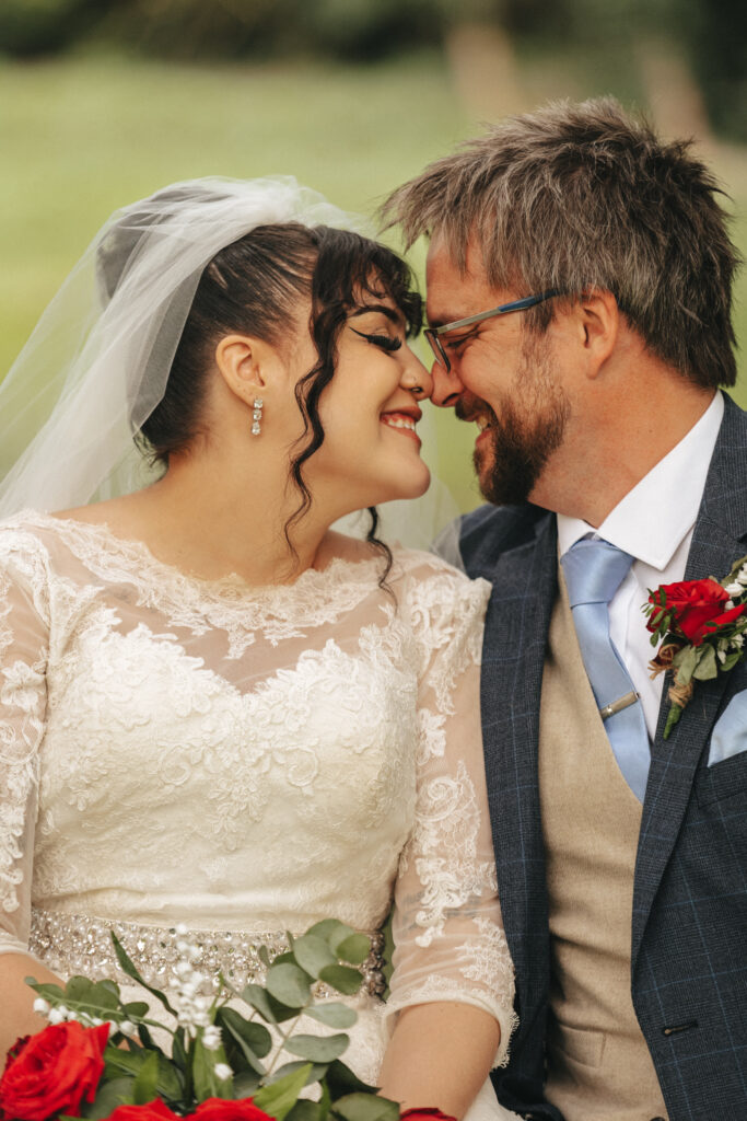 A joyful couple on their wedding day sits closely, nose-to-nose, and smiling. The bride wears a white lace gown with a veil, earrings, and a bejeweled belt, holding red roses. The groom wears glasses, a blue tie, and a gray vest under a blue suit, with a red rose boutonniere. © Aimee Lince Photography