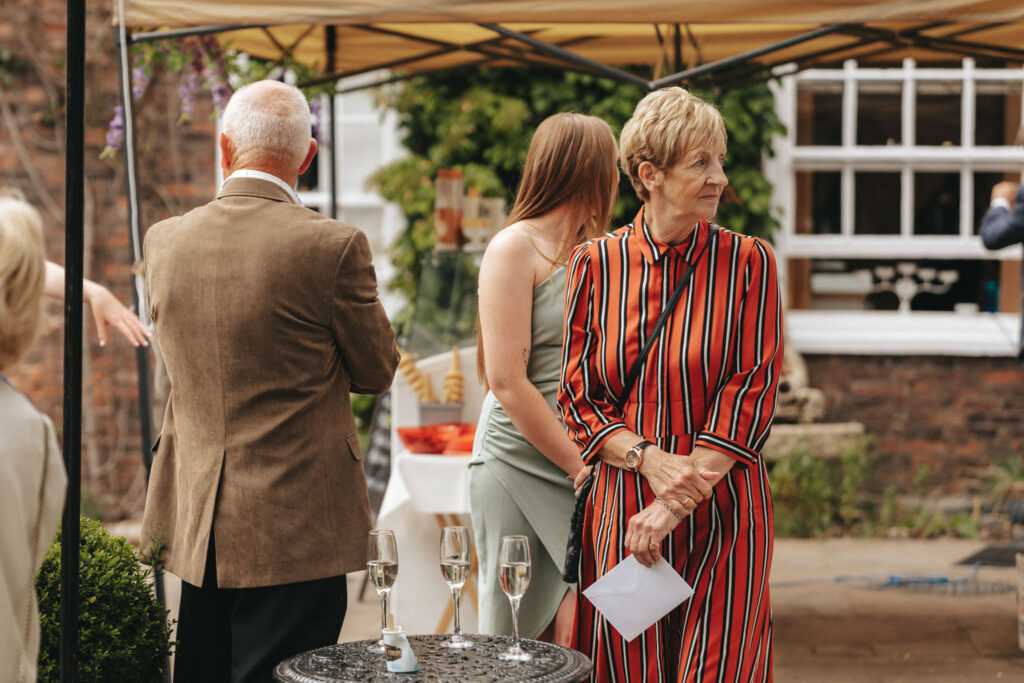 At a picturesque Bardney Hall wedding, an elderly woman in a colorful striped dress holds a note beside a young woman in light green. Both face away from the photographer capturing the moment, while a man in a brown jacket stands near. A table with champagne glasses rests under a canopy against the brick backdrop. © Aimee Lince Photography