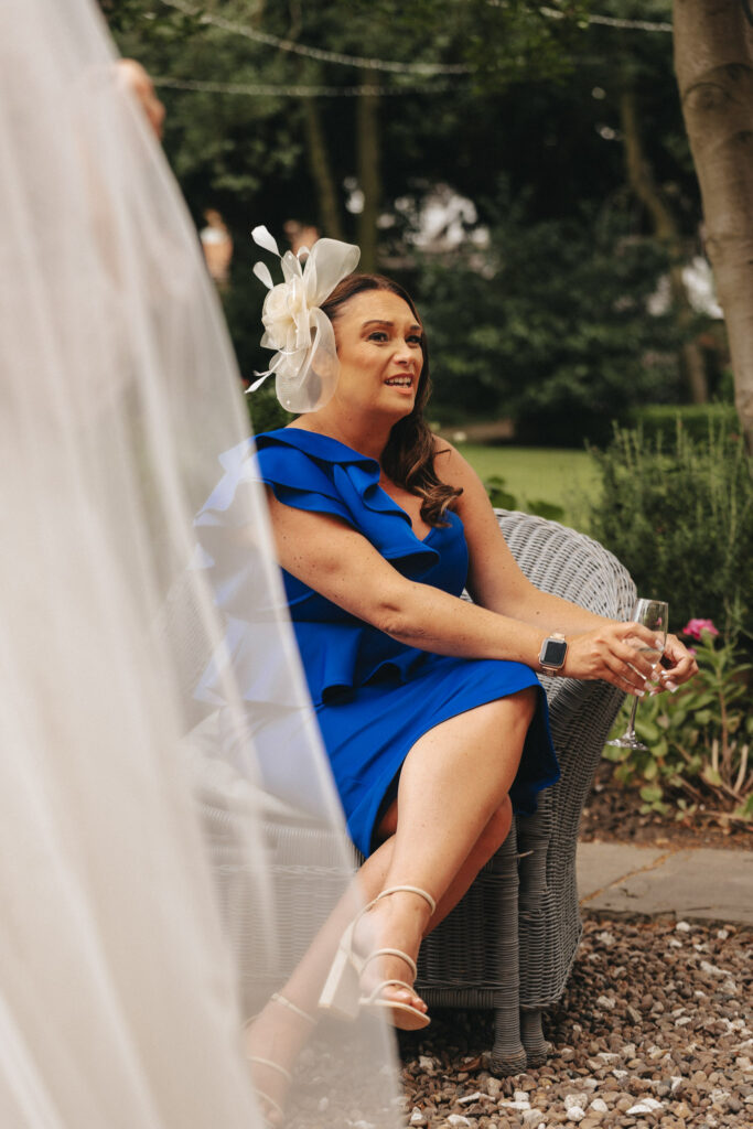 A woman in a bright blue, ruffled one-shoulder dress sits on a wicker chair outdoors at Bardney Hall, holding a champagne flute. With long brown hair and a large white fascinator, she complements the lush greenery backdrop. A bridal veil is visible on the left, capturing the photographer's eye. © Aimee Lince Photography