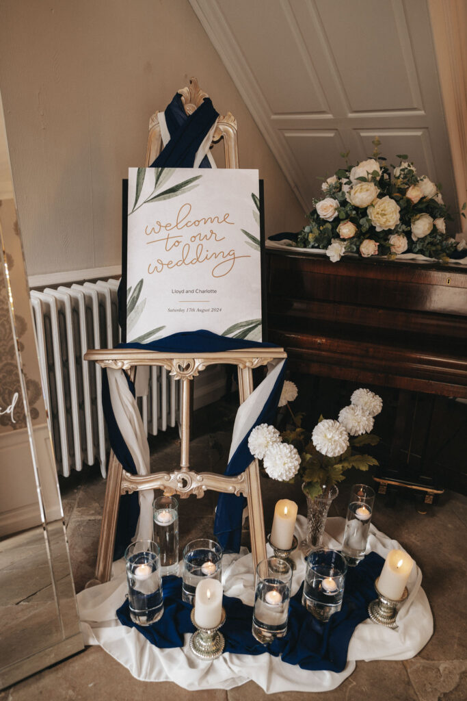 A wedding welcome sign on a wooden easel, elegantly scripted with "Welcome to our wedding" and names below. This Bardney Hall setup features navy and white fabric, surrounded by white flowers, candles in glass holders, and a bouquet atop a dark piano in the background—perfect for photography. © Aimee Lince Photography