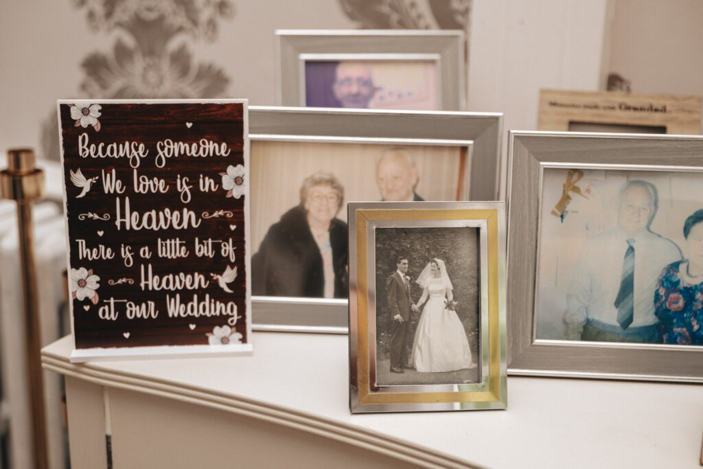 A collection of framed photographs on a shelf, captured by a talented photographer, includes a black-and-white wedding photo, colorful family portraits, and a decorative sign. The sign reads, "Because someone we love is in Heaven, there is a little bit of Heaven at our Wedding. © Aimee Lince Photography