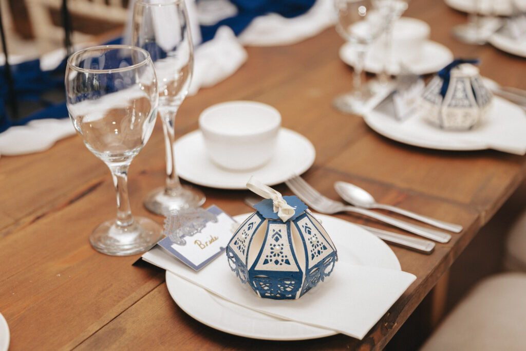 A wooden table is elegantly set for a formal meal at Bardney Hall, featuring two wine glasses, a white bowl, and a decorative blue-and-white lantern. Silver cutlery is neatly arranged with a "Bride" name card visible. White and blue tablecloth accents enhance the setting, perfect for photography. © Aimee Lince Photography
