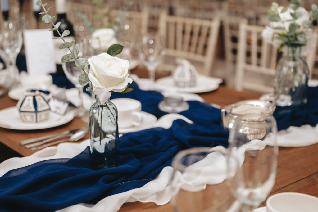 A wooden table at Bardney Hall is elegantly set with a flowing blue and white fabric runner. Blue glass vases hold white roses and eucalyptus, perfect for wedding photography. The table features fine glassware, teacups, and silver cutlery with small patterned favor boxes by each setting. © Aimee Lince Photography