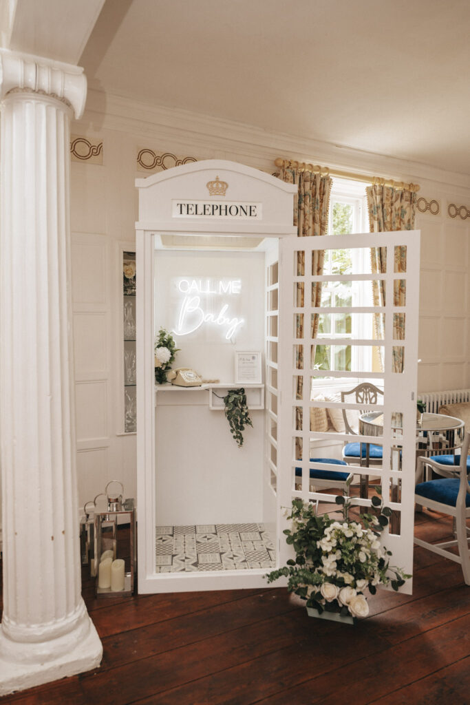 A vintage white telephone booth at Bardney Hall, adorned with a "CALL ME Baby" neon sign inside, makes for an enchanting wedding photography spot. A rotary phone on a small shelf sits amid decorative flowers, with natural light streaming in and a large white column enhancing the elegant surroundings. © Aimee Lince Photography