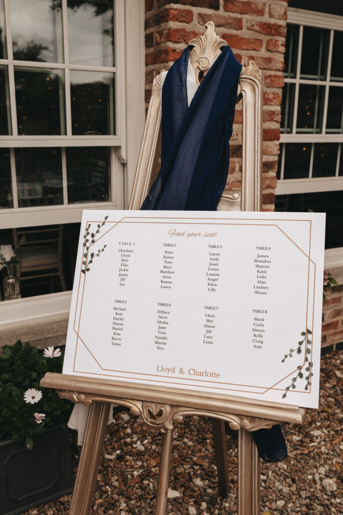 A wedding seating chart elegantly positioned on a gold easel at Bardney Hall features a white board listing tables and guest names. The "Find your seat" heading is adorned with decorative foliage, while dark blue and white draped fabric adds charm. A rustic brick backdrop completes the picturesque setting for any photographer. © Aimee Lince Photography