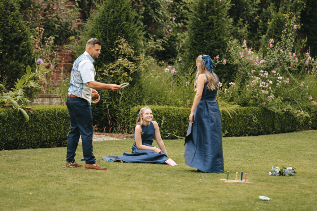 A man and two girls enjoy a lively game of ring toss on the lush lawn at Bardney Hall. The man, poised with a ring, is dressed in blue like the smiling girls. Hedges and flowering plants create a picturesque backdrop perfect for capturing this delightful moment through photography. © Aimee Lince Photography