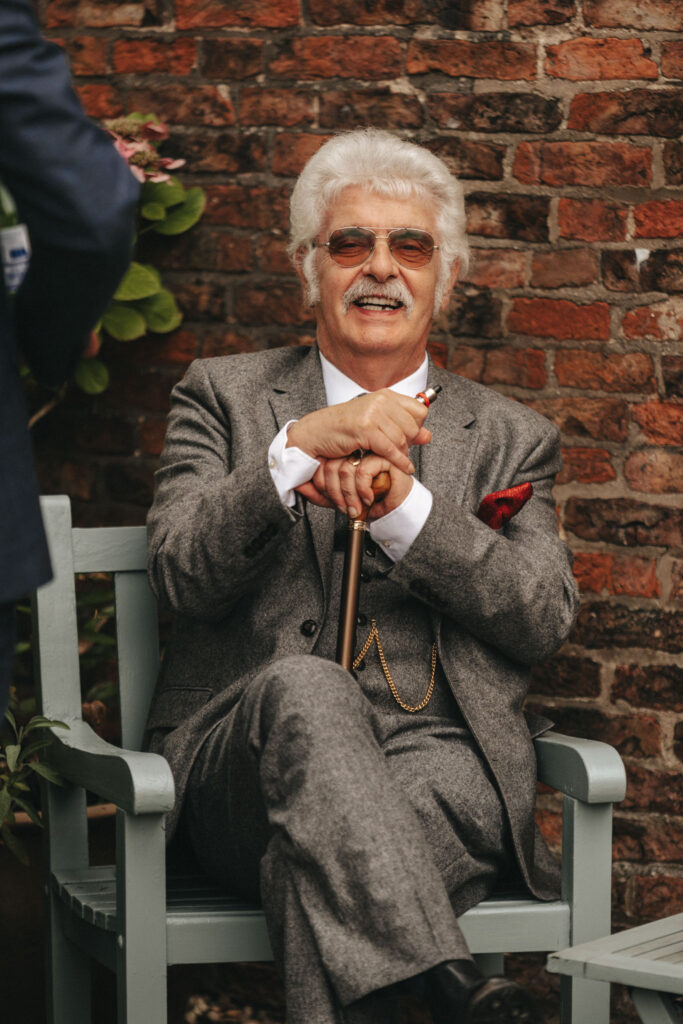 An elderly man with white hair and mustache sits on a bench, elegantly poised for a photograph. Dressed in a gray suit, white shirt, and red pocket square, he holds a wooden cane. A gold chain adorns his vest. The rustic brick wall backdrop with green leaves adds charm to the photography setting. © Aimee Lince Photography