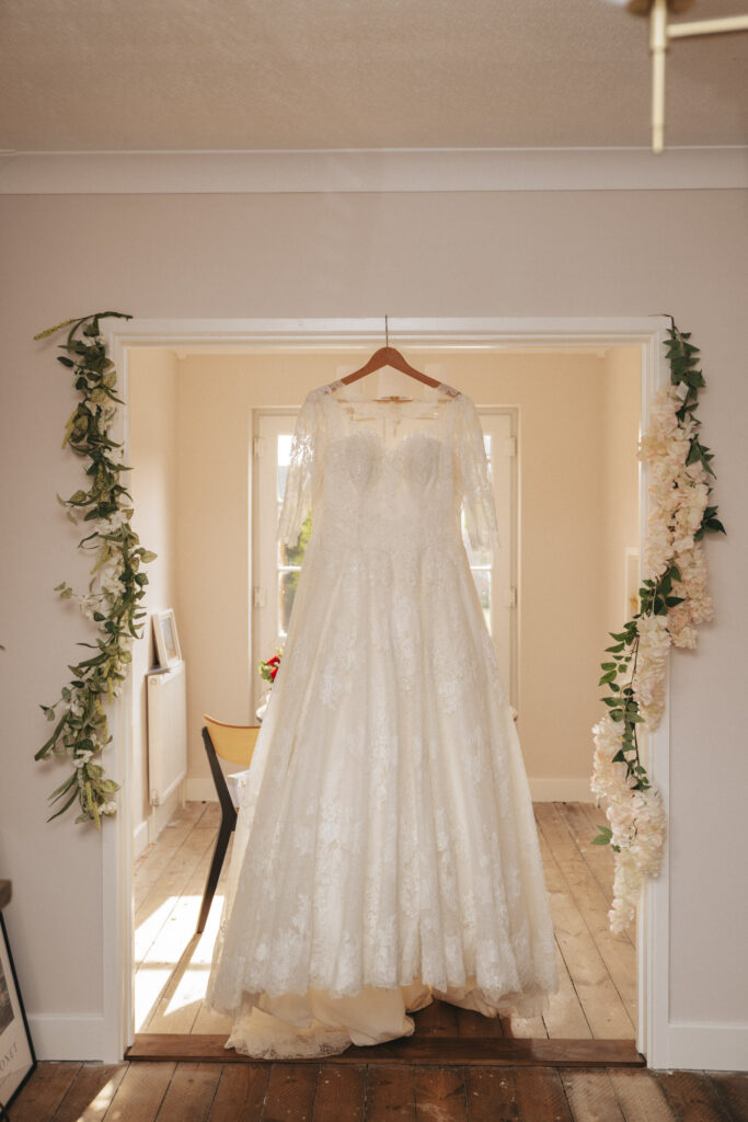 A lace wedding dress with three-quarter sleeves hangs from a wooden hanger in a bright room at Bardney Hall. The backdrop features light walls and hardwood floors, as floral garlands of white and light pink flowers drape around the doorway, capturing the perfect scene for any photographer. © Aimee Lince Photography