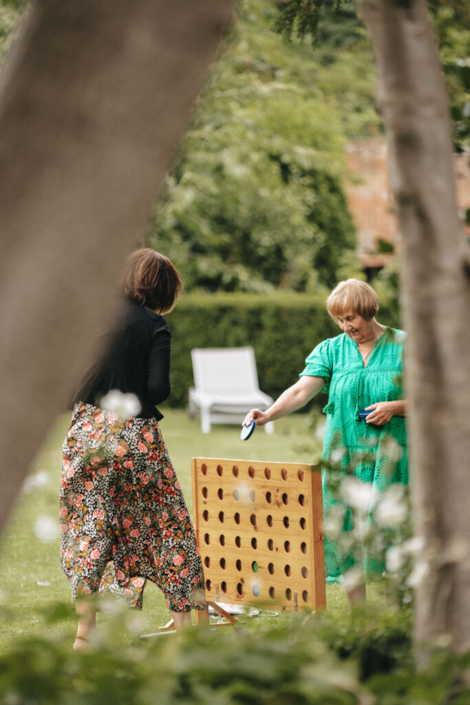 Two women, perhaps guests at a Bardney Hall wedding, play a giant Connect Four game on the lawn. One wears a floral skirt and black top, the other a green dress. They both hold game pieces, while a photographer captures the scene amid white lounge chairs and greenery framed by tree trunks. © Aimee Lince Photography
