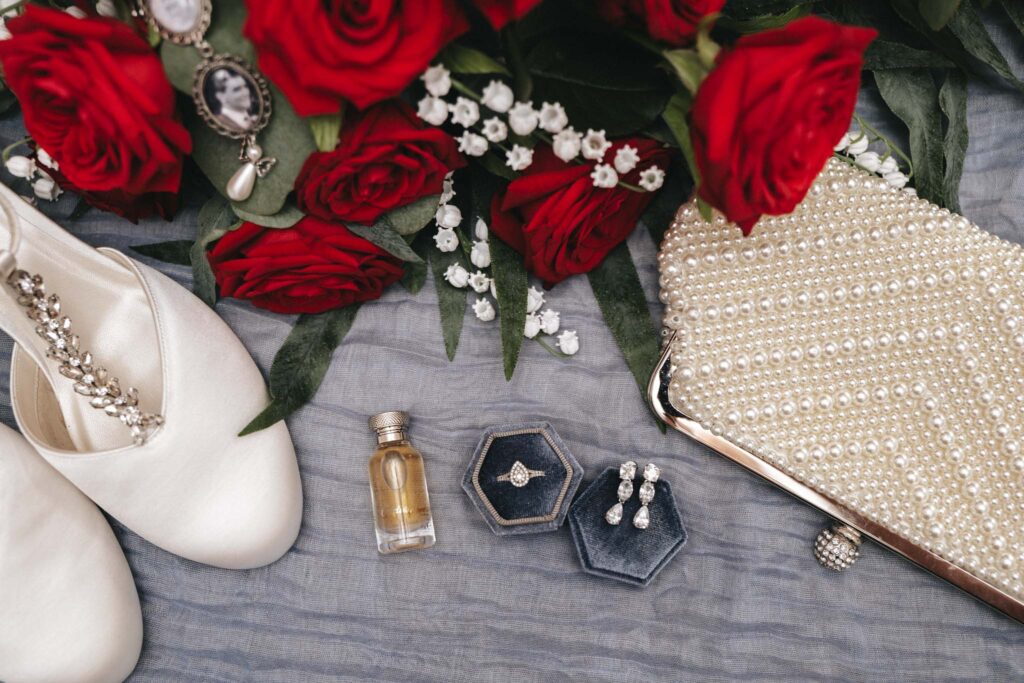 A flat lay captures the essence of a romantic wedding at Bardney Hall, featuring red roses with white baby's breath, a pearl-studded clutch, elegant white shoes with jeweled straps, a small perfume bottle, vintage cameo earrings, and jewelry boxes containing a ring and pearl earrings on crumpled gray fabric. © Aimee Lince Photography