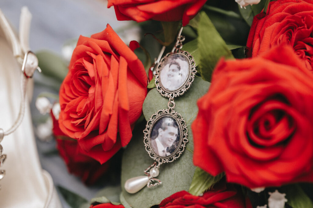 A close-up of vibrant red roses with green leaves showcases a vintage-style locket adorned with sepia-toned portraits. One locket dangles a teardrop pearl and heart charm, creating a scene suggestive of romance and nostalgia, enhanced by bridal shoes nearby—a perfect wedding capture at Bardney Hall. © Aimee Lince Photography