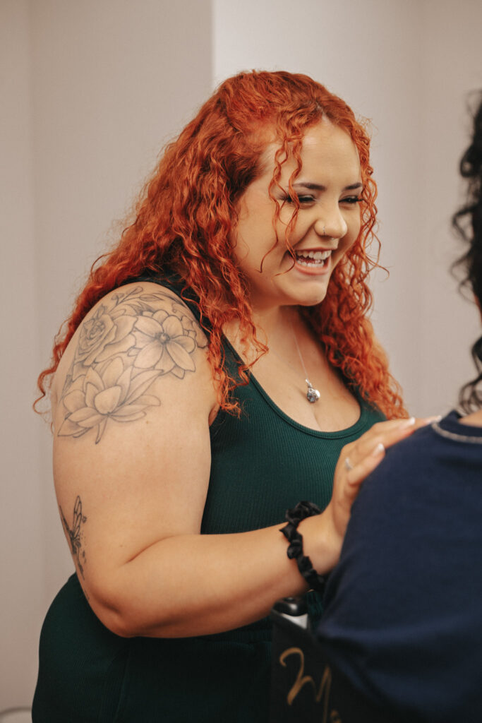 Smiling woman with long curly red hair and a floral tattoo on her upper arm stands in a dark green tank top. She holds hands with a person partially visible in the foreground, wearing a dark shirt. Captured by a talented photographer, the backdrop is a plain, light-colored wall. © Aimee Lince Photography
