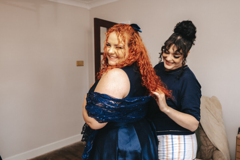 Two smiling women are preparing for an event indoors at Bardney Hall. The woman in front, with long curly red hair, wears an off-shoulder dark blue gown. The woman behind, with curly black hair in a bun, helps adjust the dress as they both cheerfully engage in the fitting process before the photographer arrives. © Aimee Lince Photography