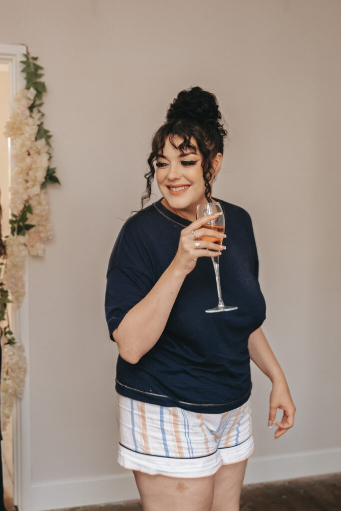 A woman with dark hair in a bun, clad in a navy t-shirt and striped shorts, holds a glass of wine, smiling and gazing to the side. She stands indoors at Bardney Hall against a plain wall, with flowers visible in the background near an open door—a moment worthy of a photographer's eye. © Aimee Lince Photography