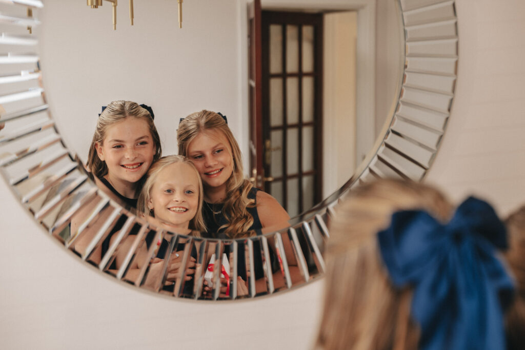 Seen through a decorative round mirror, three girls with light hair and cheerful smiles brighten the warmly lit room at Bardney Hall. One, adorned with a blue bow, adds charm to their ensemble of dark tops. The mirror's silver frame, featuring a spoke-like design, enhances this delightful wedding photography moment. © Aimee Lince Photography