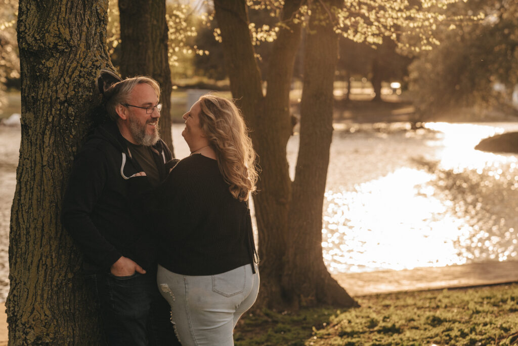 A couple enjoys a moment together by a serene lake during sunset. The man, wearing a hoodie and jeans, leans against a tree smiling, while the woman, in a black sweater and light blue jeans, stands close to him, also smiling. The sun casts a warm glow, reflecting off the water in the background. © Aimee Lince Photography