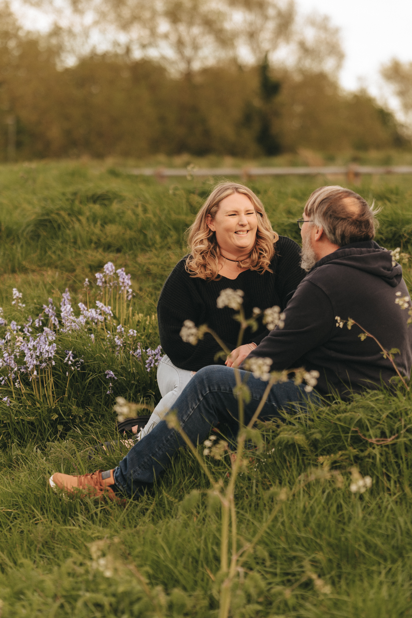 A couple sits on a grassy field, smiling at each other. The woman has blonde hair, and the man has gray hair and a gray beard. The woman is wearing a black sweater, and the man is wearing a black hoodie. There are blooming blue flowers nearby, and trees are visible in the background. © Aimee Lince Photography