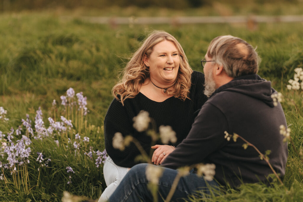 A woman and a man sit on a grassy field surrounded by wildflowers at Cleethorpes Boating Lake. They are facing each other and smiling warmly. The woman has long, wavy hair and is wearing a black sweater. The man has short hair, a beard, glasses, and is wearing a dark hooded sweatshirt. A blurred fence and greenery are in the background. © Aimee Lince Photography