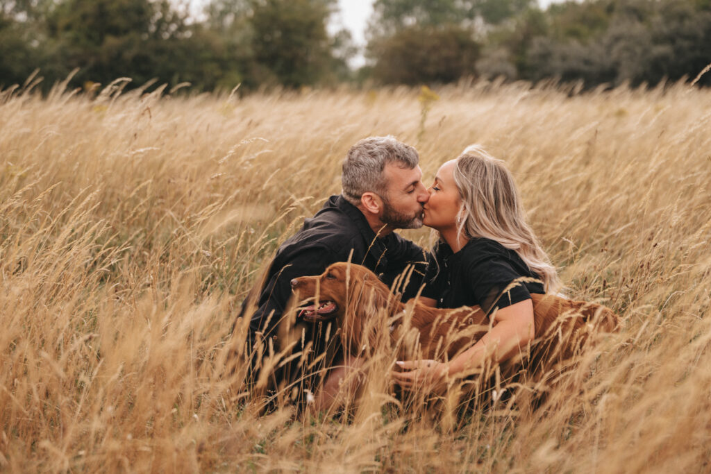 A couple with a dog share a kiss while kneeling in a field of tall, golden grass at Cleethorpes Beach. Both are dressed in black clothing. The man has short gray hair and the woman has long blond hair. The brown dog is sitting between them, panting contentedly. Trees and a cloudy sky are visible in the background. © Aimee Lince Photography