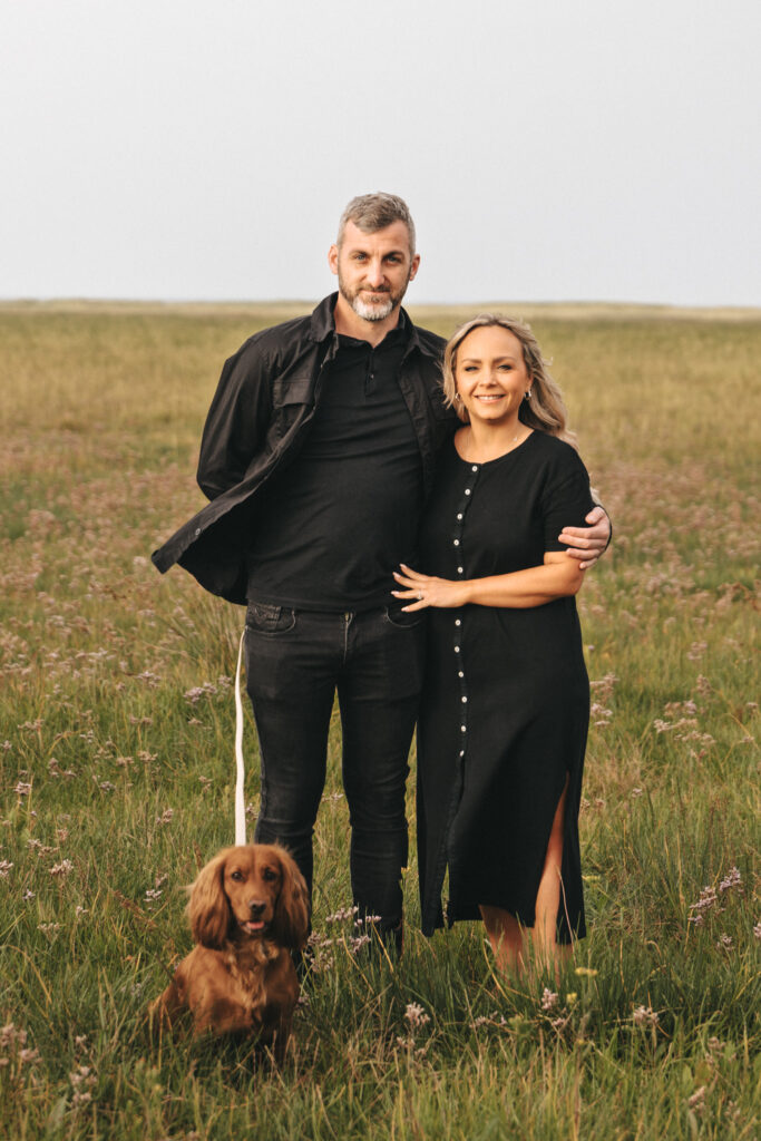 A man and woman stand together in a grassy field, both dressed in black. The man is wearing a jacket and jeans, and the woman is in a button-down dress with a side slit. They are smiling and the man has his arm around the woman. A small brown dog sits at their feet, looking at the camera. © Aimee Lince Photography