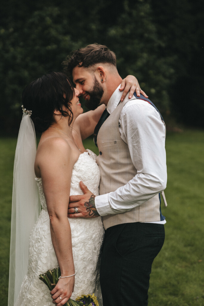 A bride and groom share an intimate moment outdoors. The bride, in a white lace gown and veil, holds a bouquet of flowers and embraces the groom with her right hand. The groom, wearing a white shirt, tie, and beige vest, with tattoos visible on his left hand, gently touches the bride's back. © Aimee Lince Photography