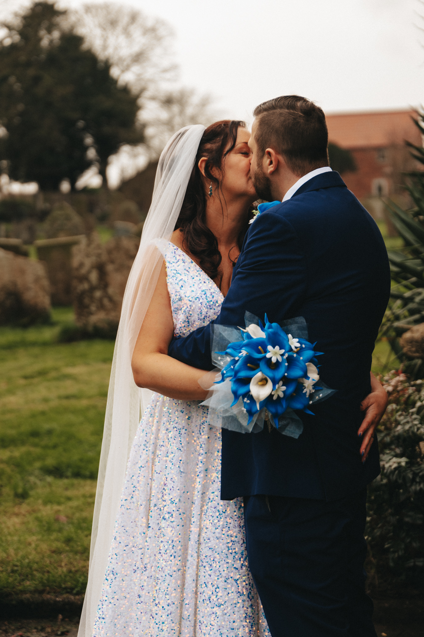 A bride and groom share a kiss outdoors. The bride, dressed in a white gown with shimmering details, holds a bouquet of white and blue flowers. The groom wears a dark blue suit. They stand on a green lawn with trees and an old building in the background. The bride's hand rests on the groom's back. © Aimee Lince Photography