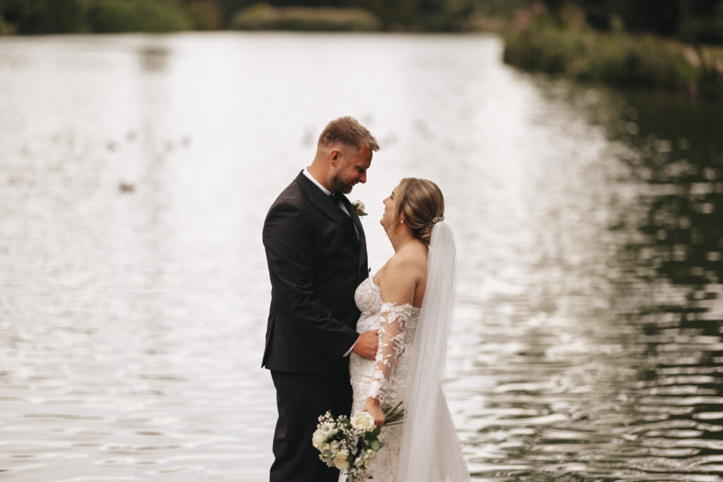 A bride and groom stand close together sharing a joyful moment by the serene lake at Elsham Hall. The groom wears a black suit with a white shirt, and the bride is in a lacy, off-shoulder wedding dress. She holds a bouquet, and both smile warmly at each other against the backdrop of calm water and greenery. © Aimee Lince Photography