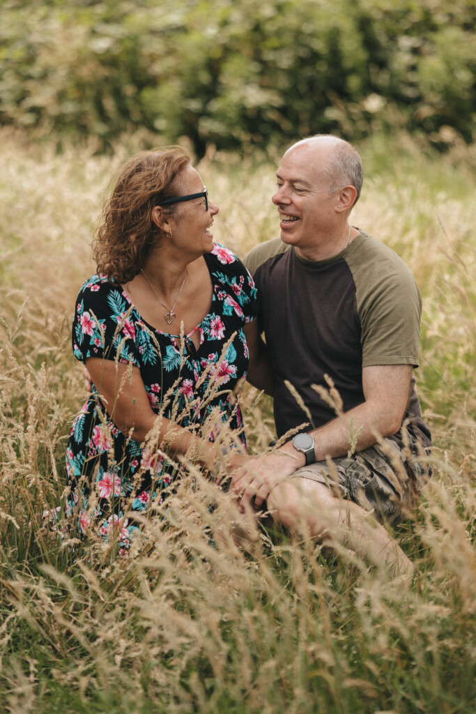 A couple sits in a grassy field, smiling and looking at each other. The woman wears a dark floral dress and glasses, while the man wears a short-sleeved, two-tone shirt and shorts. They are surrounded by tall grass, and greenery is visible in the background. © Aimee Lince Photography