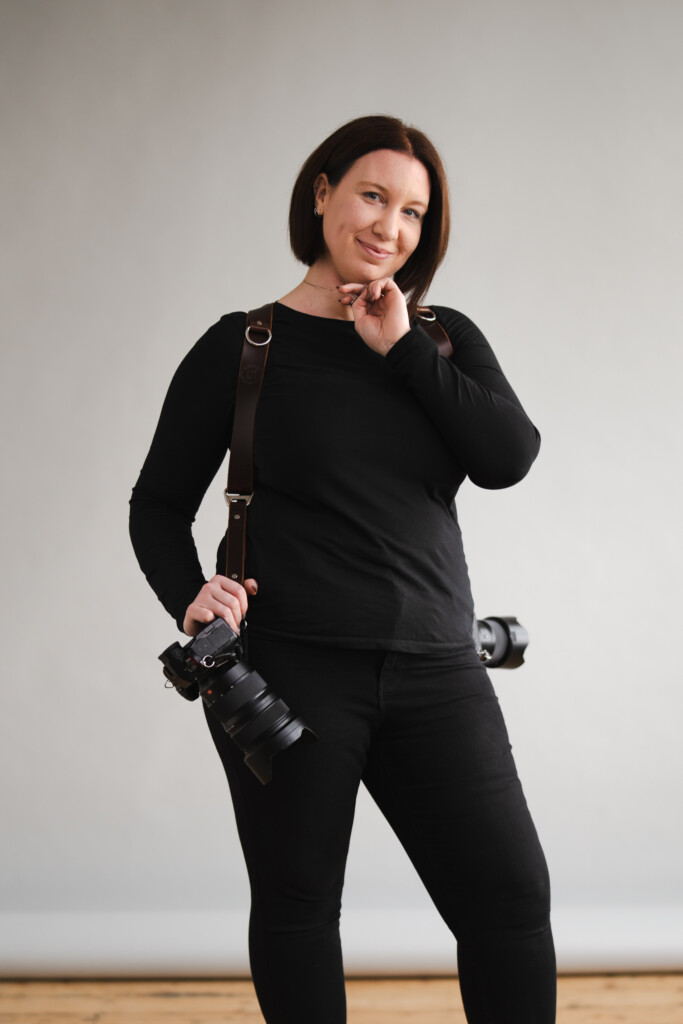 A person with short, dark hair, dressed in a black long-sleeve shirt and black pants, poses against a neutral gray background. They wear a dual camera strap harness holding two cameras, one in each hand, while resting their right hand on their chin and slightly smiling at the camera. © Aimee Lince Photography