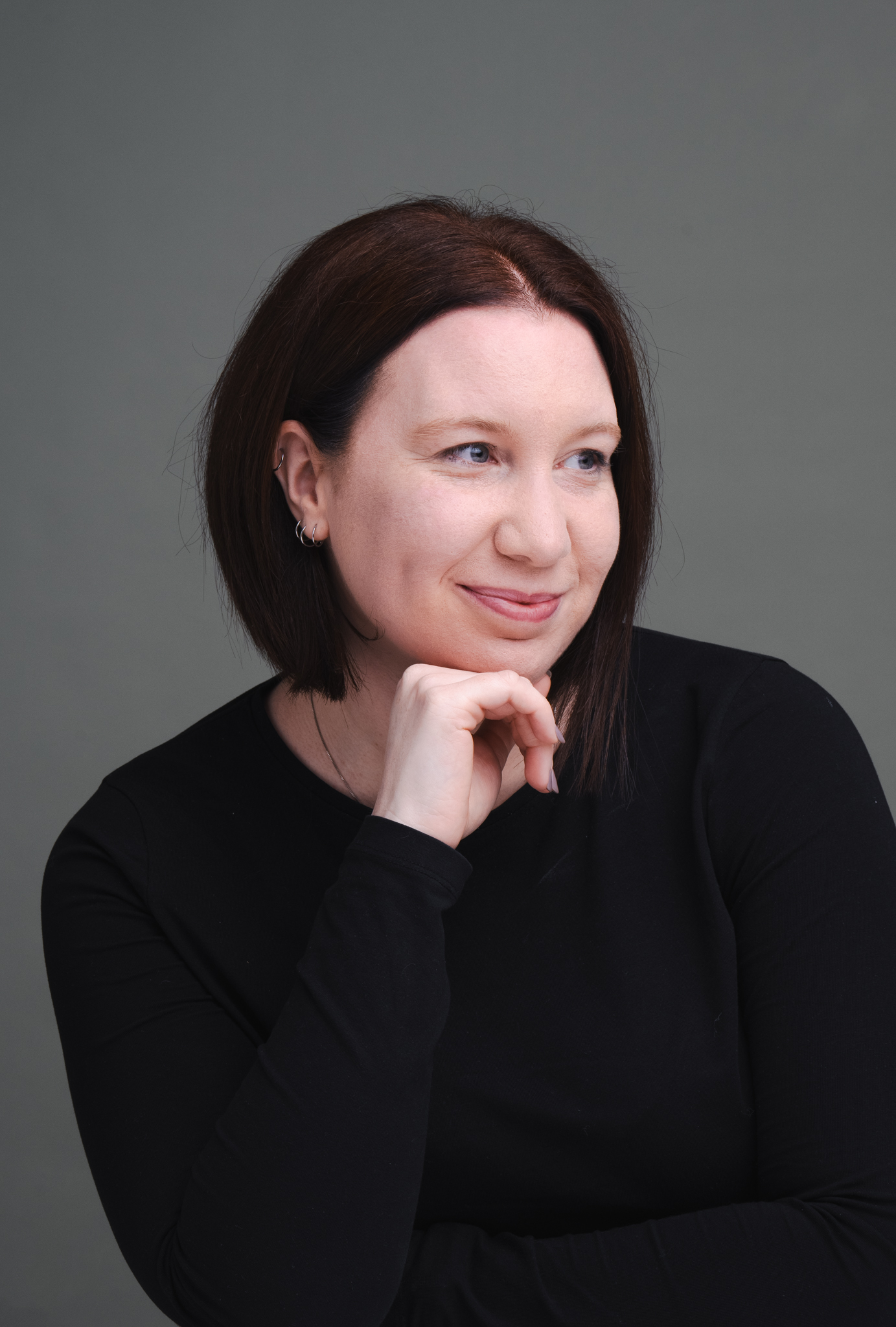 A person with shoulder-length dark hair, wearing a long-sleeved black shirt, and small hoop earrings. They are smiling slightly and resting their chin on their right hand with a grey background. © Aimee Lince Photography