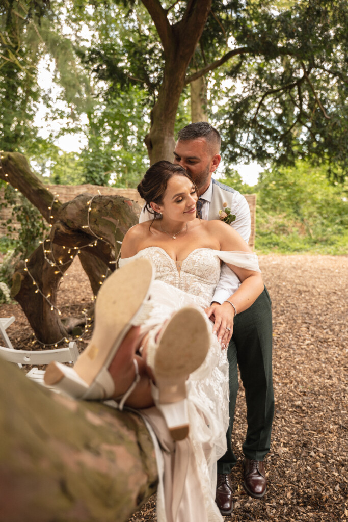 A bride and groom sit in a nature setting. The bride, in a sleeveless lace wedding dress, reclines playfully with her feet up on a tree branch, showing her sandals. The groom, in dark trousers and a white shirt, sits behind her, leaning in for a kiss. The scene is lit with soft, natural light. © Aimee Lince Photography