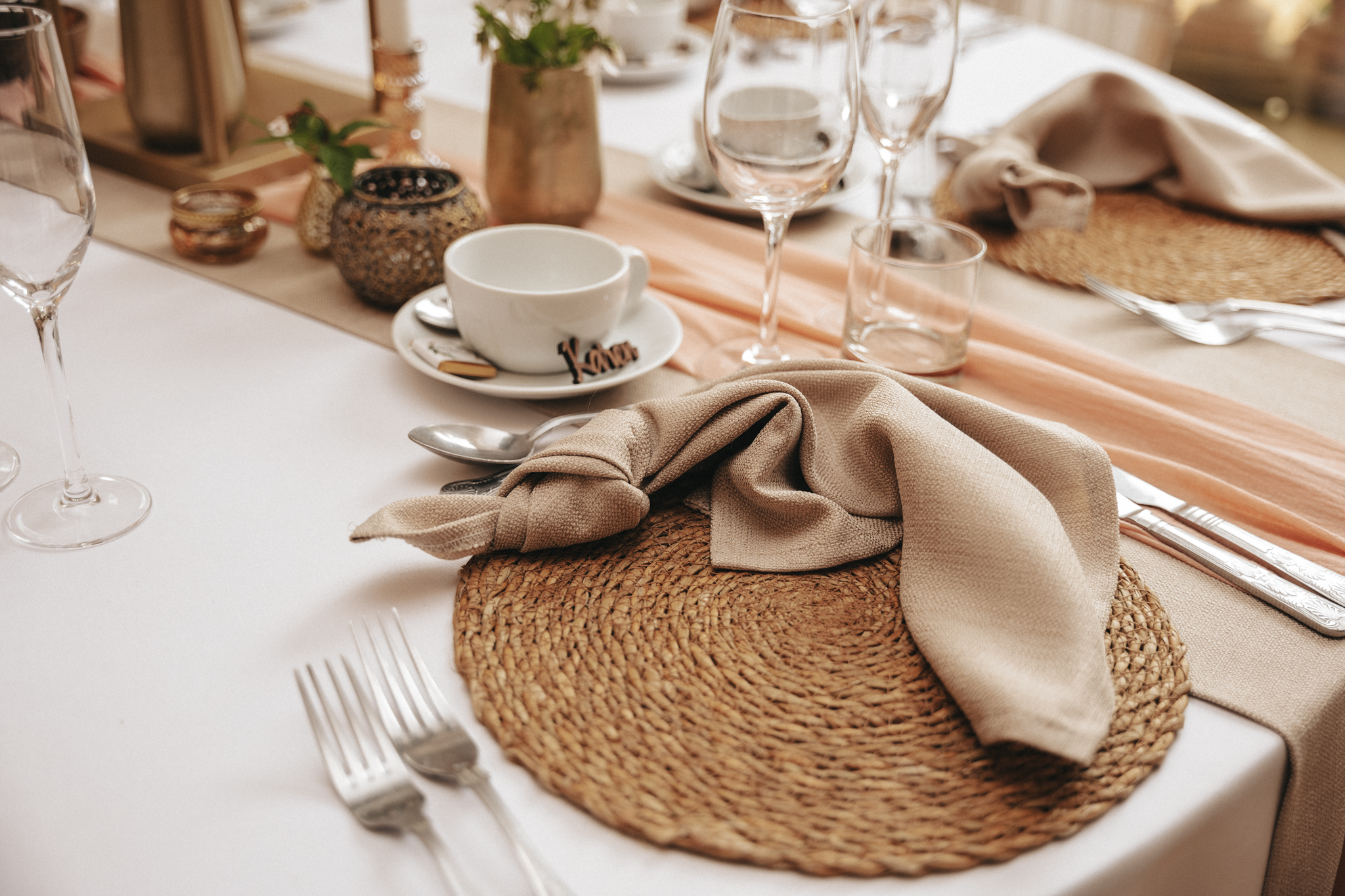 An elegant table setting features a woven placemat, silverware, a beige cloth napkin stylishly draped, a white cup and saucer with a nametag reading "Kara," several glassware items, and decorative elements like small candles and greenery, all arranged neatly and tastefully for a formal event. © Aimee Lince Photography