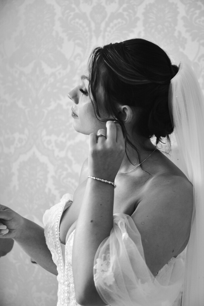 Black and white photo of a bride adjusting her earring. She is wearing a strapless, lace wedding gown with a sweetheart neckline and sheer sleeves. Her hair is styled in an elegant updo, adorned with a long, flowing veil. The background features a subtle, ornate damask pattern. © Aimee Lince Photography