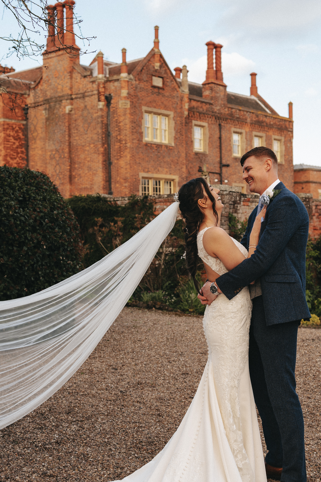 A bride in a white dress with a long veil and a groom in a blue suit are embracing and smiling at each other in front of a large, old brick building with multiple chimneys and windows. The setting appears to be an outdoor garden area with hedges and gravel pathways. © Aimee Lince Photography