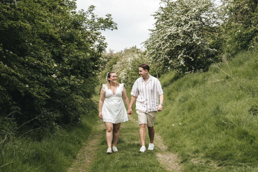 A couple walks hand-in-hand down a grassy, tree-lined path on a sunny day. The woman, wearing a white dress, smiles at the man, who is dressed in a striped shirt and beige shorts. Both are wearing white shoes. Flowering trees and lush greenery surround them, indicating a vibrant, nature-filled setting at Irby Dales Wood. © Aimee Lince Photography