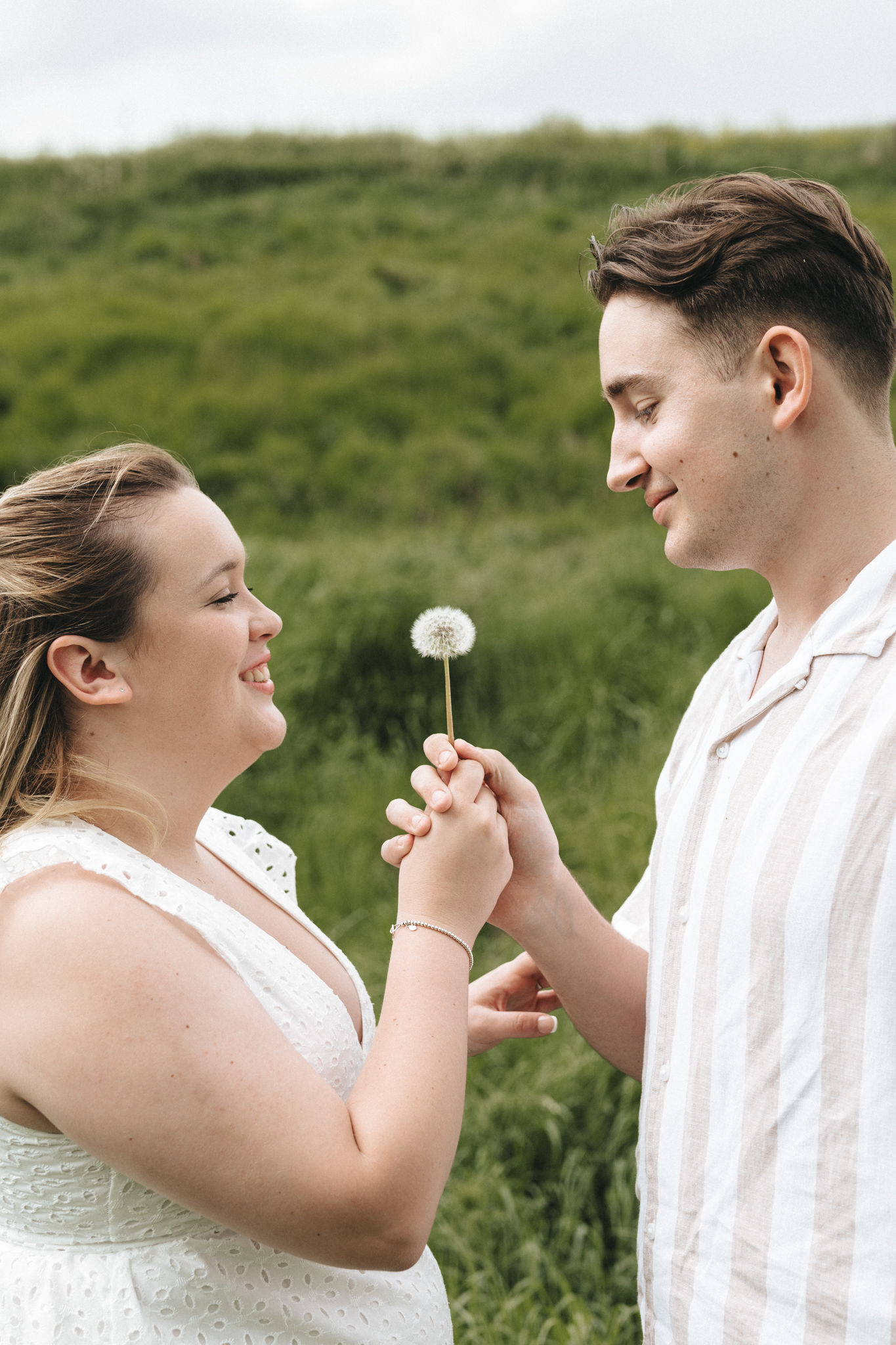 A woman and a man in a grassy field smile at each other while holding a dandelion together. The woman, on the left, wears a white, sleeveless dress; the man, on the right, in a striped shirt. They share a moment of joy, with greenery filling the background. © Aimee Lince Photography