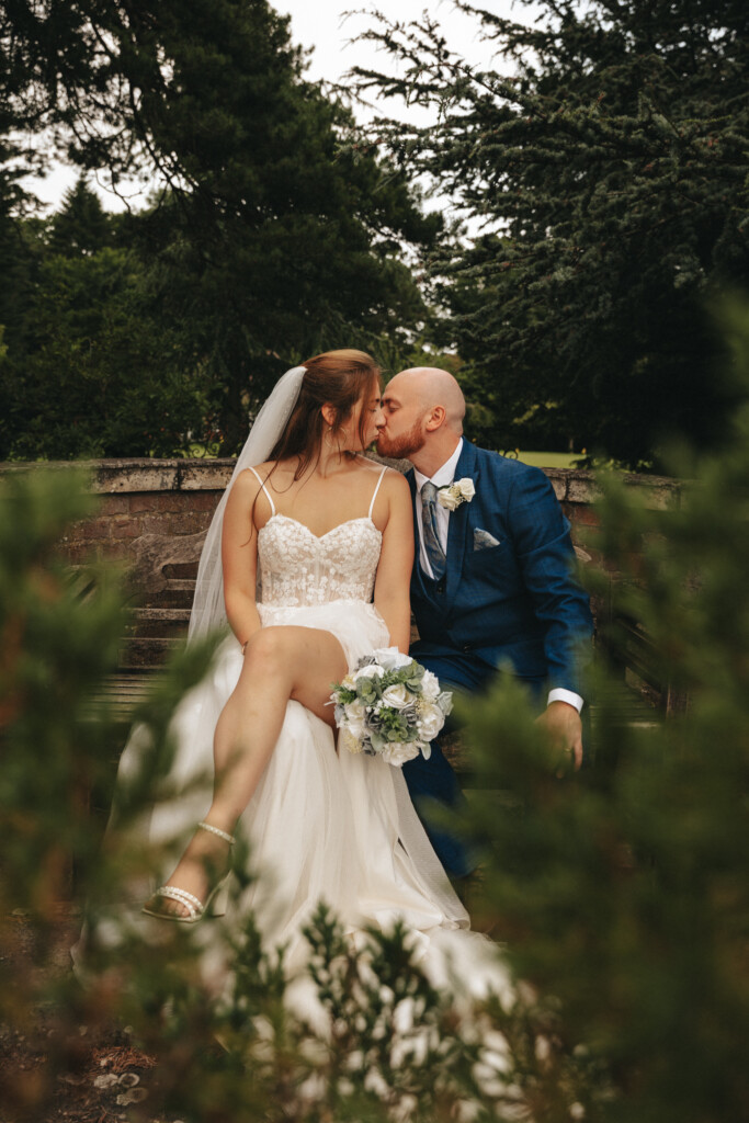 A bride and groom share a kiss while sitting on a wooden bench outdoors. The bride wears a strapless, lace-bodied gown with a tulle skirt and holds a bouquet of white and blue flowers. The groom is dressed in a blue suit and white shirt. Greenery surrounds them, creating a romantic atmosphere. © Aimee Lince Photography