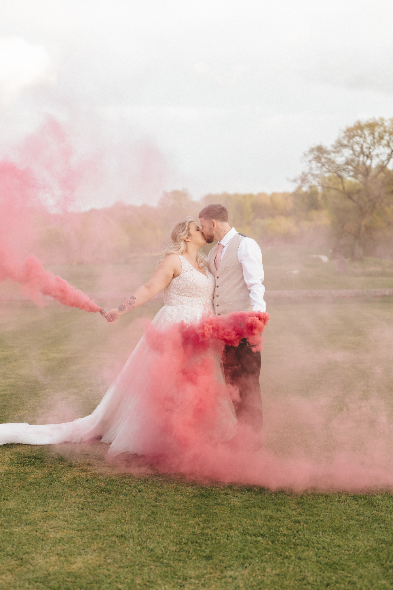 A bride in a white gown and a groom in a white shirt and beige vest kiss while holding smoke bombs emitting pink smoke outdoors on a grassy field. The bride's dress has a long train, and trees are visible in the background on a slightly cloudy day. © Aimee Lince Photography