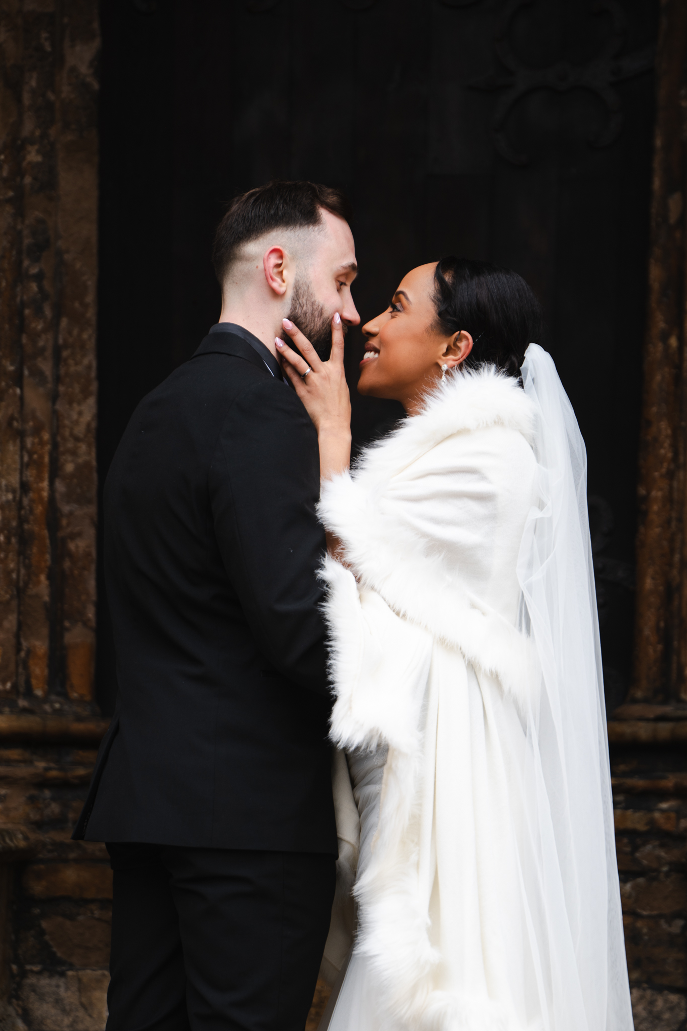 A bride and groom lovingly gaze into each other's eyes. The bride, wrapped in a white fur-trimmed cloak and veil, gently touches the groom's face. The groom, dressed in a black suit, holds her waist. They stand in front of a textured, dark background, possibly an ornate wooden door. © Aimee Lince Photography