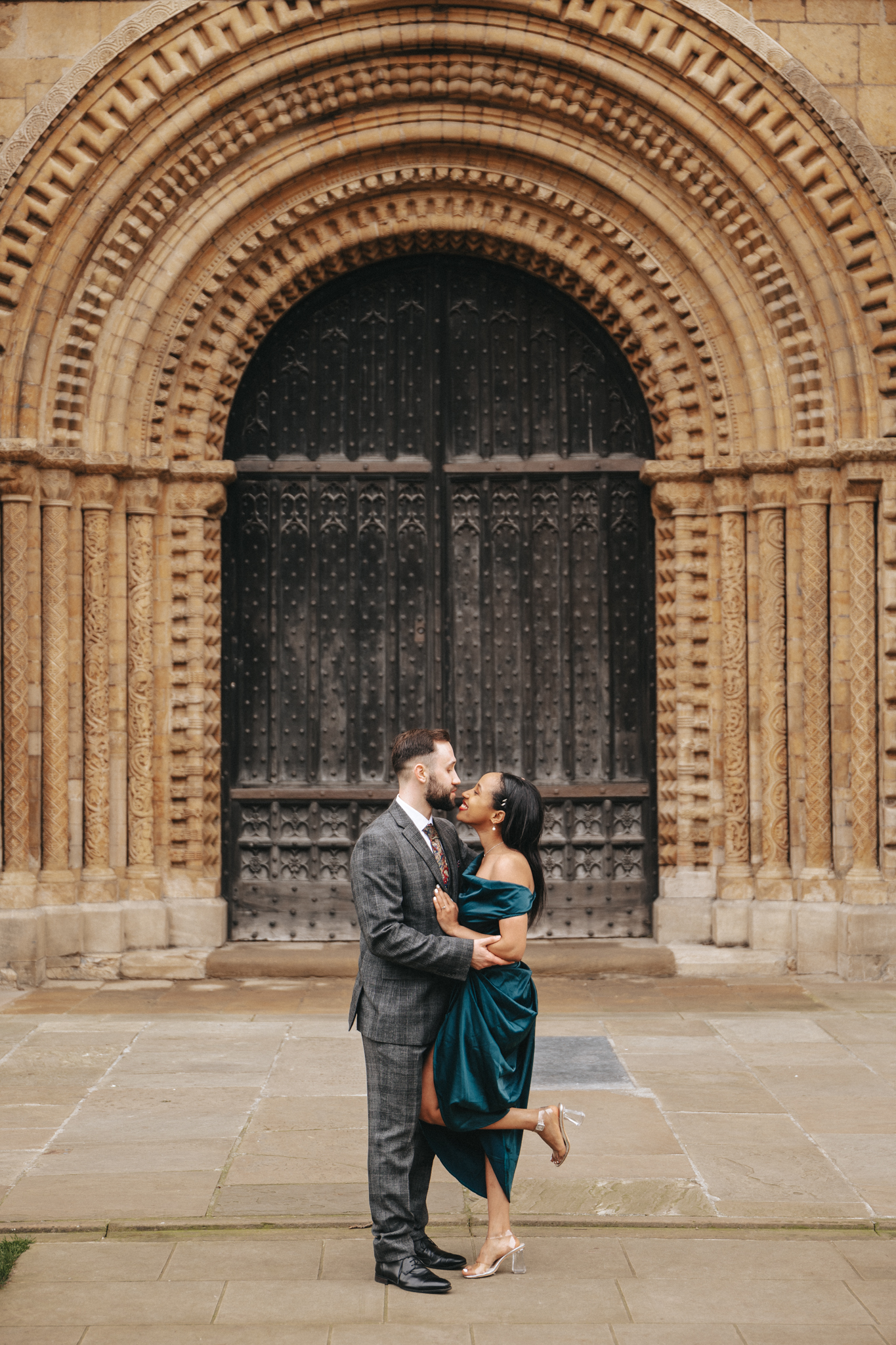 A couple stands in front of a grand, intricately patterned arched doorway made of stone. The woman, wearing a teal dress and heels, gazes up at the man, who is dressed in a plaid suit and tie. They hold each other close on a stone walkway, surrounded by the detailed architecture. © Aimee Lince Photography