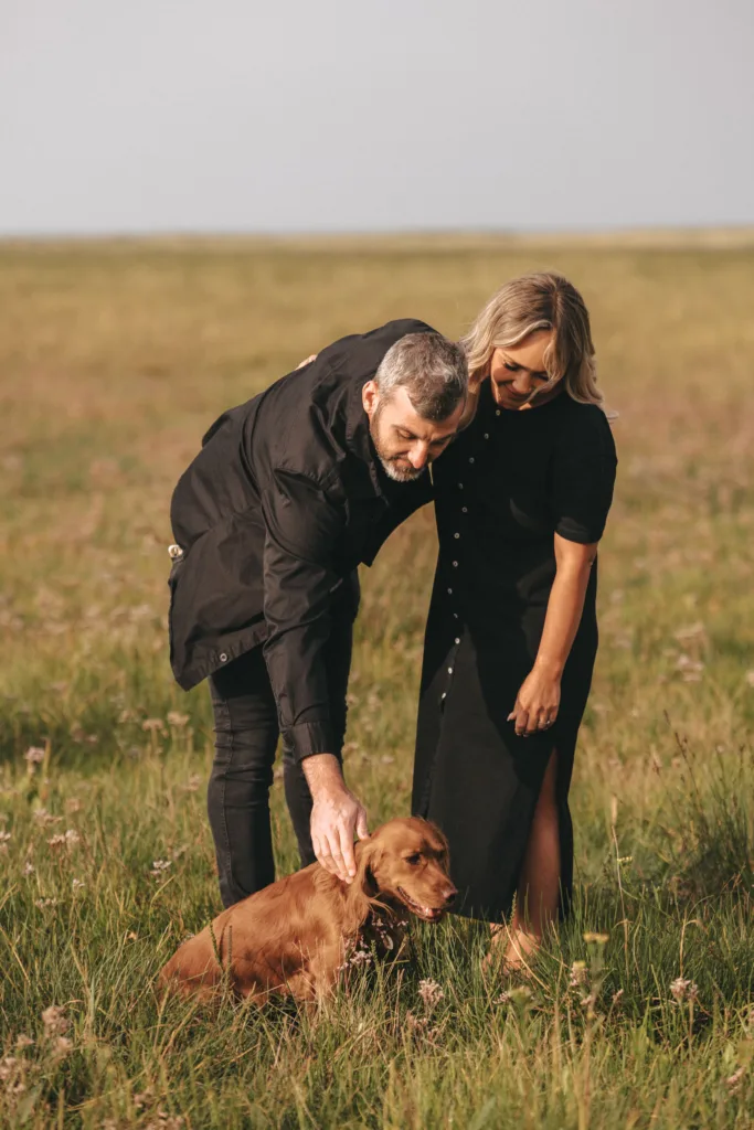 A man and woman, both in black outfits, stand in a grassy field under a clear sky. They are bending down to gently pet a brown dog sitting on the ground. Recently engaged, the couple appears relaxed and content, sharing this tranquil moment with their dog in the natural landscape. © Aimee Lince Photography - Wedding photographer in Lincolnshire, Yorkshire & Nottinghamshire