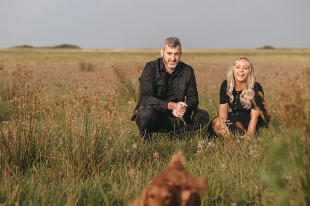 A man and woman crouch in a field of tall grass, both clad in black outfits. The man has short hair and a beard, while the woman, with long blonde locks, is smiling as their brown dog sprints towards them. With an overcast sky overhead, it feels like the perfect moment for an engagement at Cleethorpes Beach. © Aimee Lince Photography - Wedding photographer in Lincolnshire, Yorkshire & Nottinghamshire