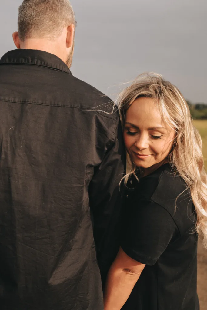 A woman with long blonde hair affectionately leans her cheek against a man's black jacket, smiling softly. The man, in a matching black shirt, faces away as they stand outdoors near Cleethorpes Beach, with green fields blurred under a gray sky in the background. © Aimee Lince Photography - Wedding photographer in Lincolnshire, Yorkshire & Nottinghamshire