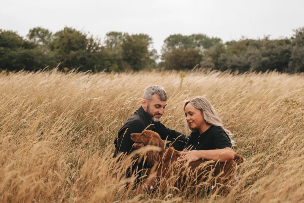 A man and woman, newly engaged, sit in a field of tall golden grass, each petting a brown dog nestled between them. Both are wearing dark clothing. Trees stand in the background under an overcast sky, adding to the serene rural setting. © Aimee Lince Photography - Wedding photographer in Lincolnshire, Yorkshire & Nottinghamshire