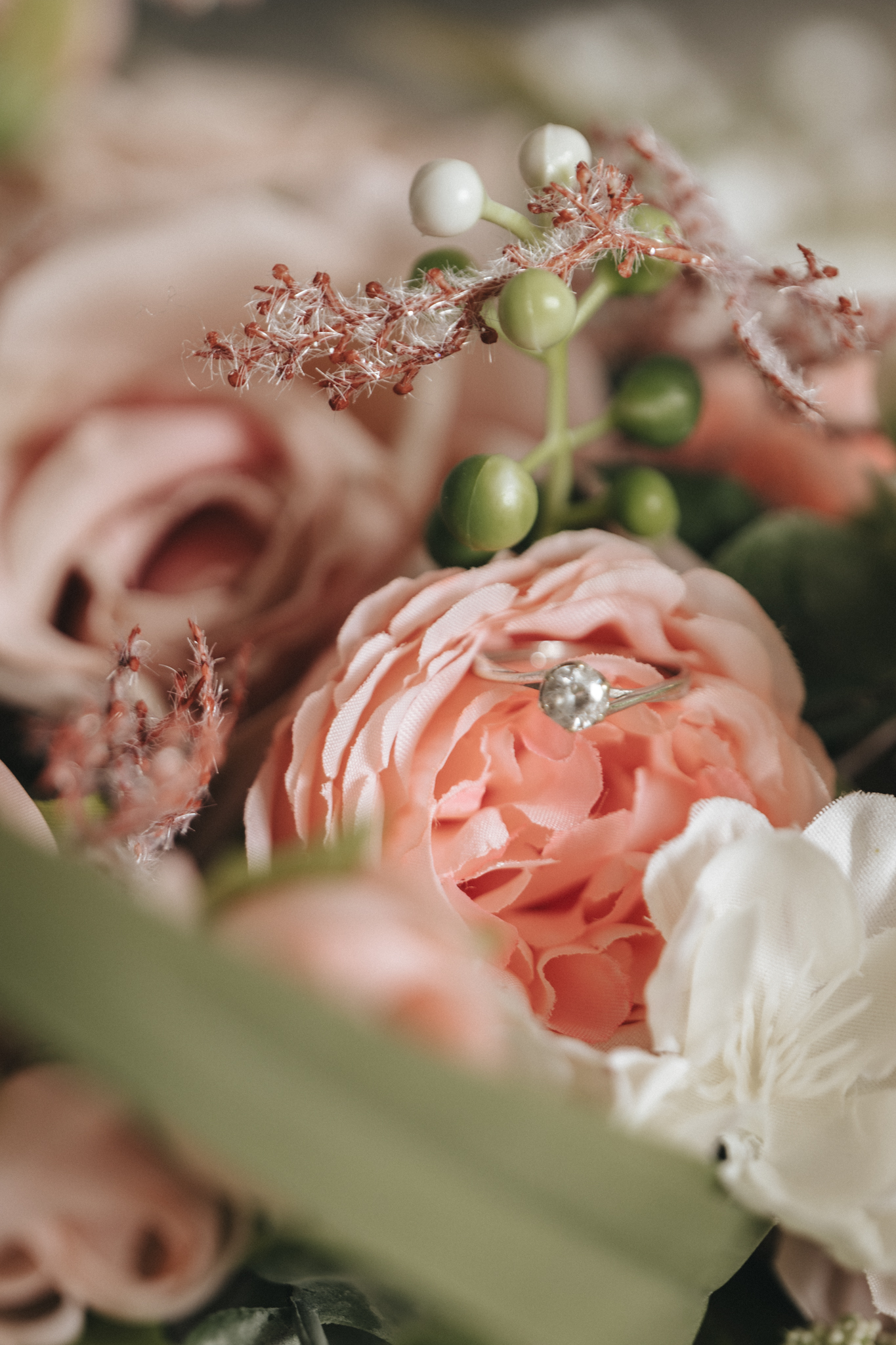 A close-up shot of a decorative bouquet featuring a prominent pink peony in the center, adorned with a diamond ring. Surrounding the peony are various other flowers and greenery, including white blossoms and green berries. Perfect for wedding photos at Oaklands Hall Hotel, this bouquet adds a touch of elegance to the scene. © Aimee Lince Photography