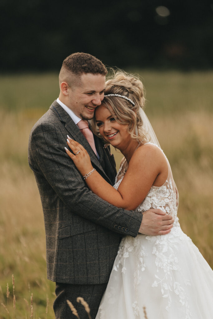 A bride and groom embrace outdoors, standing in a field of tall grass at Oaklands Hall Hotel. The bride wears a white lace wedding dress and a veil, with her hair up and a headband. The groom is dressed in a gray suit with a pink tie. Both are smiling warmly, showcasing their happiness in the wedding photos. © Aimee Lince Photography