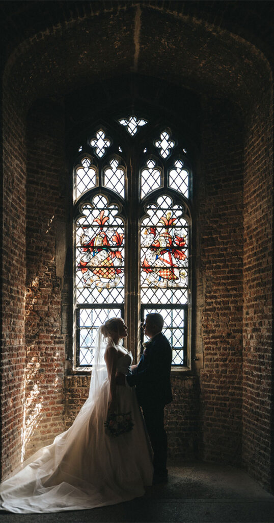 In a dimly lit, brick-walled room reminiscent of Lincolnshire charm, a bride and groom stand before an ornate stained glass window. The bride's flowing gown and veil complement the groom's suit as sunlight filters through, casting colorful patterns on the floor—a perfect scene for timeless photography. © Aimee Lince Photography